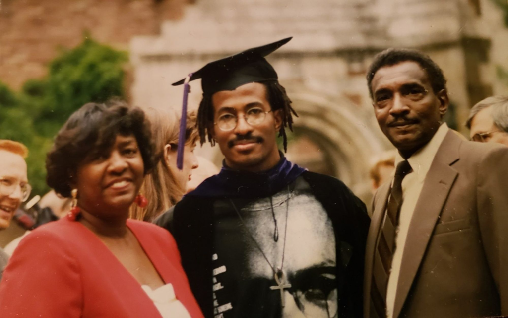 A young man wearing a graduation cap and gown stands between two adults outdoors, all smiling for a photo.