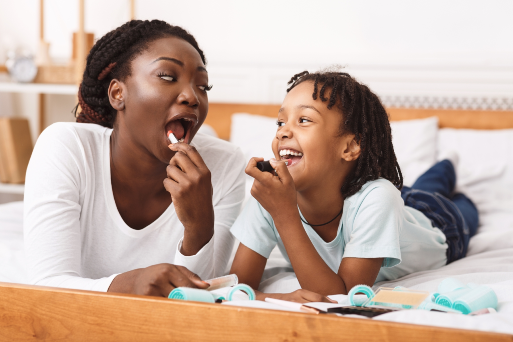 A mother and child lie on a bed smiling and applying lipstick together in a playful moment.