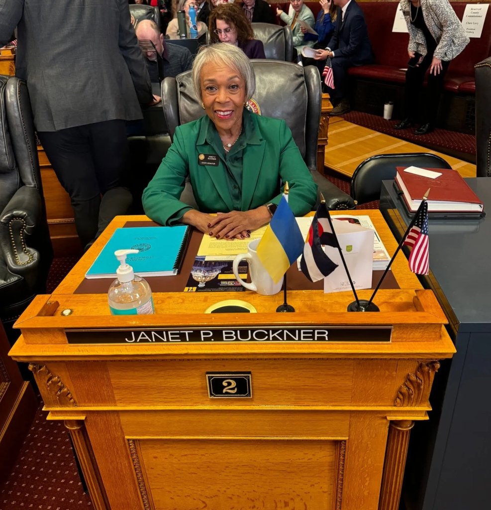 Janet Buckner sits at her desk on the Colorado Senate floor, smiling as documents and small flags rest on the desk in front of her.