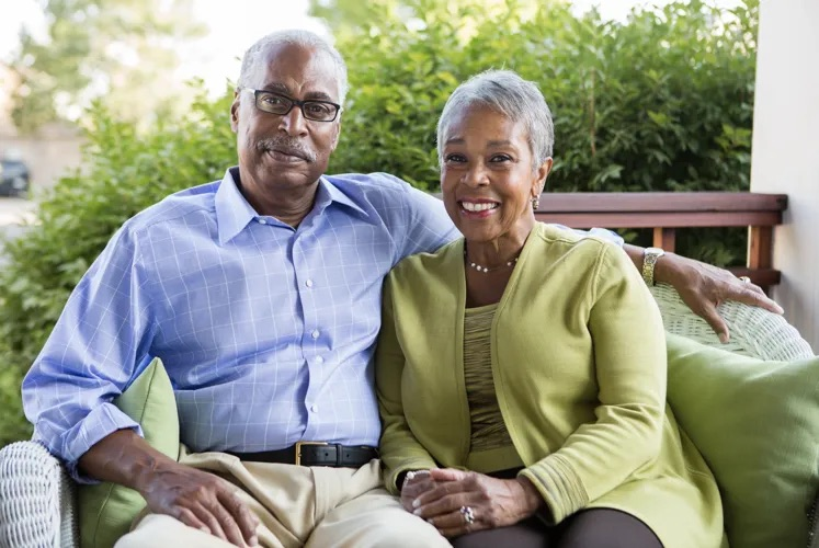 Janet Buckner sits beside her husband on an outdoor patio, both smiling as they relax together on a cushioned bench.