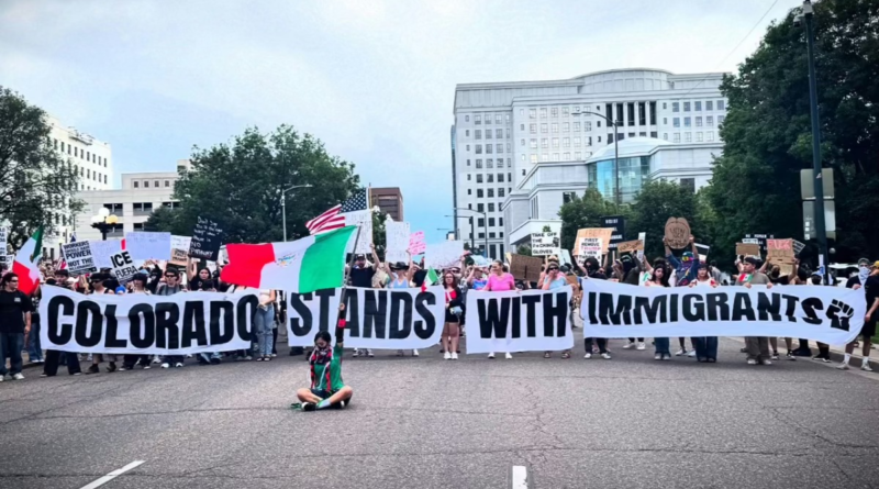 Large crowd of protesters march in downtown Denver holding a banner that reads “Colorado Stands With Immigrants” during a public demonstration.