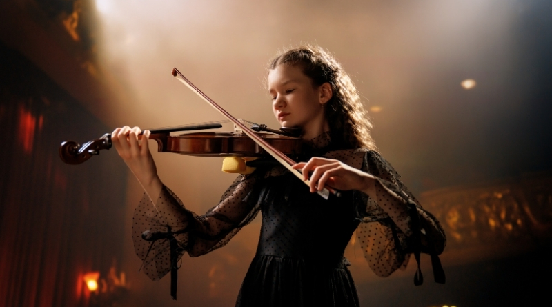 Young woman in black dress playing violin in a dark, moody setting.
