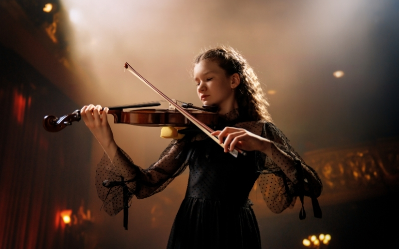 Young woman in black dress playing violin in a dark, moody setting.