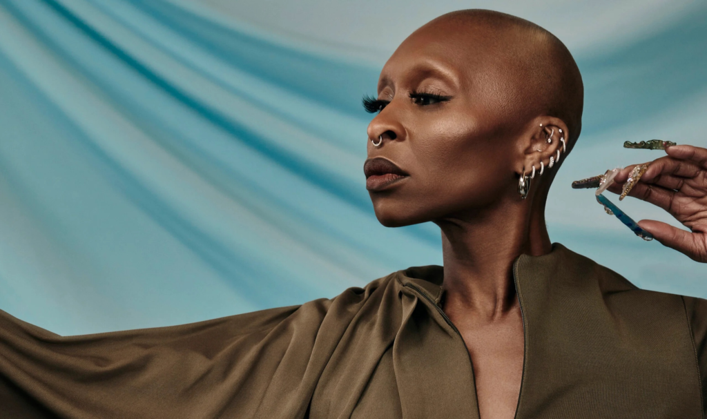 Cynthia Erivo in a stylized portrait, wearing an olive outfit with dramatic lighting, looking to the side while a hand adjusts her earring.