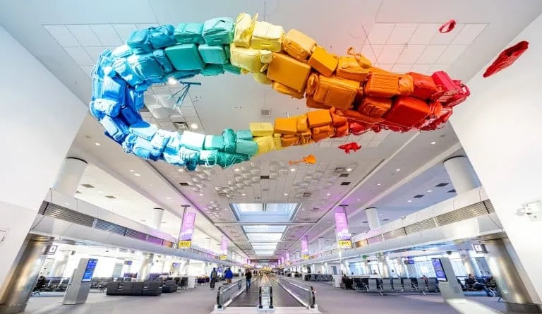 Colorful infinity-shaped art installation made of stacked suitcases hangs from the ceiling inside a spacious airport terminal.