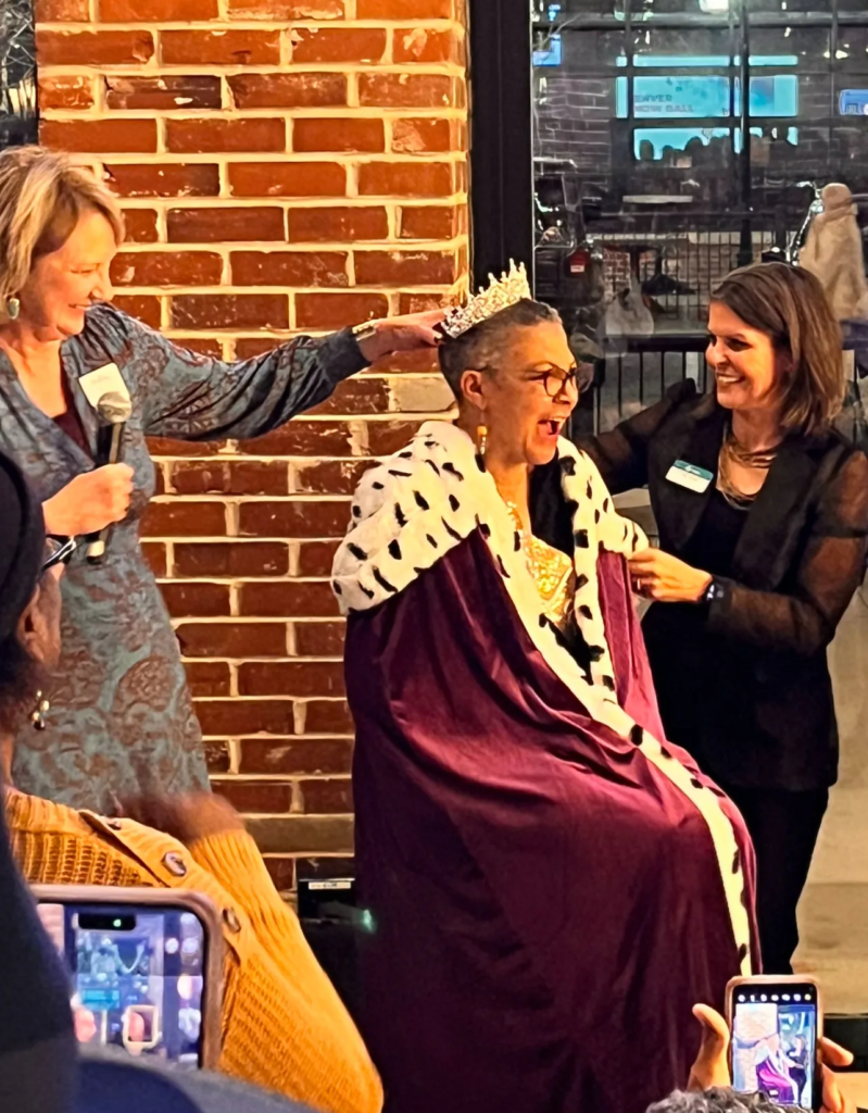 A woman reacts with surprise and joy as two people place a crown on her head during a celebratory event, with attendees capturing the moment on their phones.