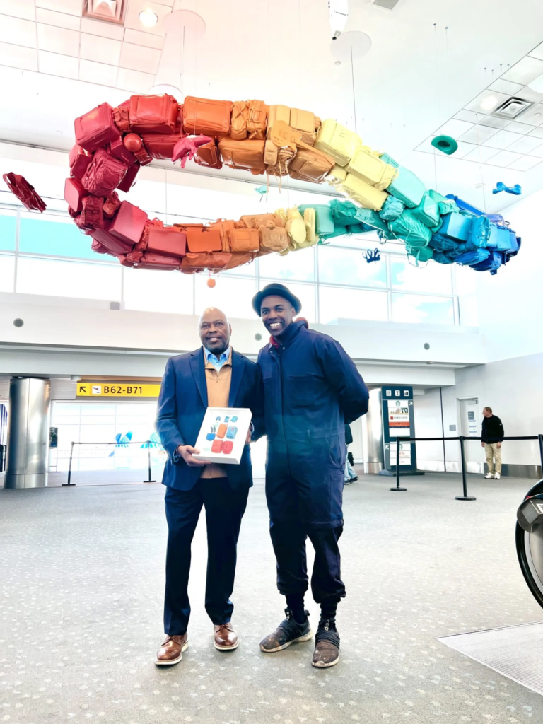 Two men stand beneath a colorful suitcase installation in an airport terminal, smiling as one holds a small artwork.