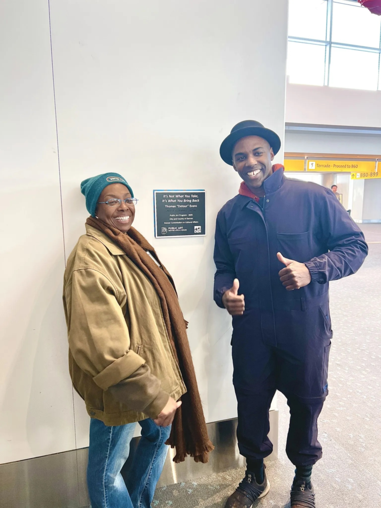 Two people stand beside a wall plaque for a public art installation in an airport, smiling as one gives a thumbs-up.