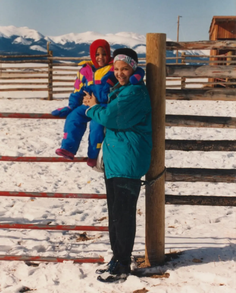 A woman in a teal jacket holds a child in colorful winter clothing while standing by a wooden fence in a snowy landscape with mountains in the background.