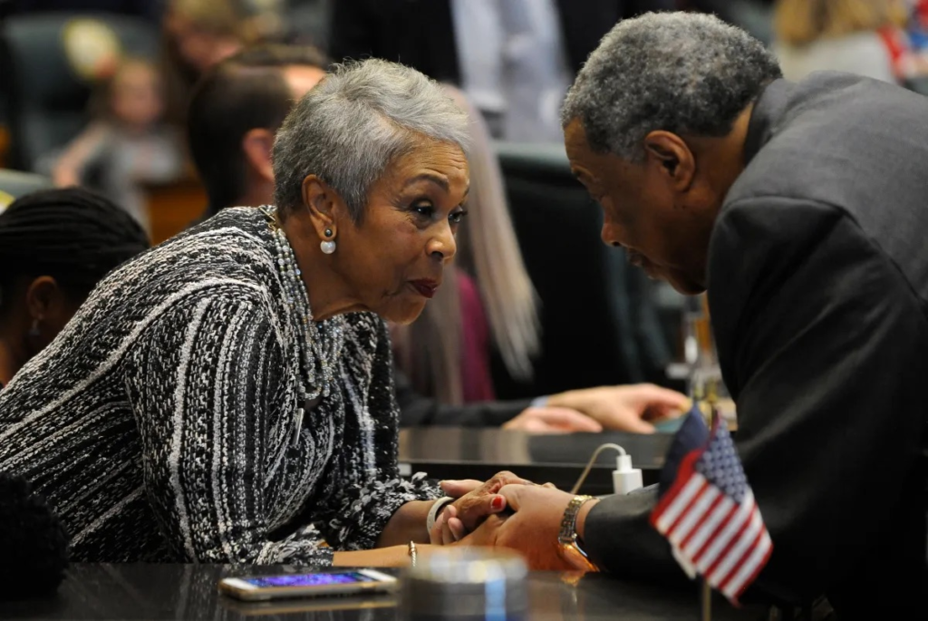 Janet Buckner leans in to speak with a colleague at the Colorado State Capitol, holding hands during a focused conversation at a legislative desk.