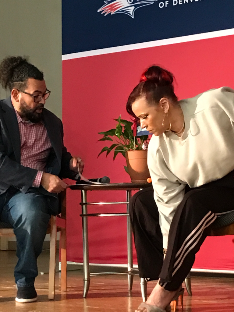 Nikole Hannah-Jones leans forward to adjust her seat during a public speaking event, while a moderator sits nearby with notes and a microphone.
