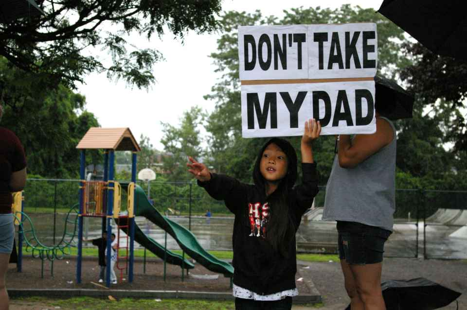 Child holding a sign that reads “Don’t Take My Dad” stands near a playground during a protest against immigration enforcement.