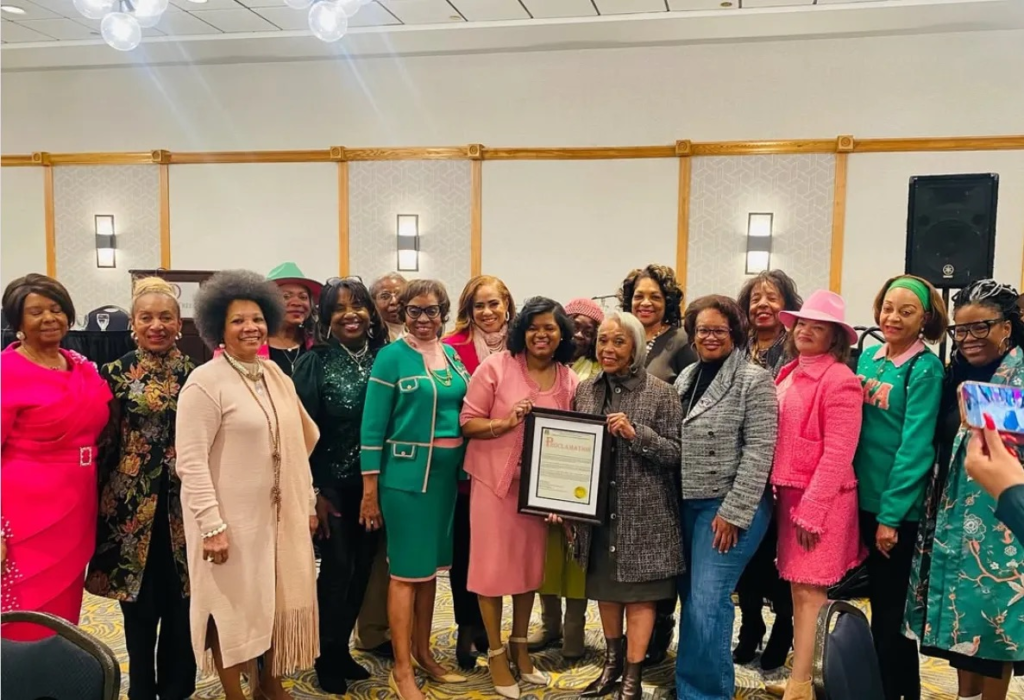 Group of women gathered indoors for a community celebration, posing together as one holds a framed proclamation honoring Janet Buckner.