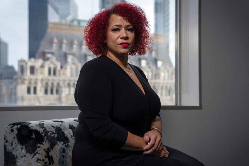 Nikole Hannah-Jones sits indoors in front of a window with a cityscape in the background, wearing a black dress and looking directly at the camera.