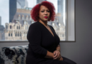 Nikole Hannah-Jones sits indoors in front of a window with a cityscape in the background, wearing a black dress and looking directly at the camera.