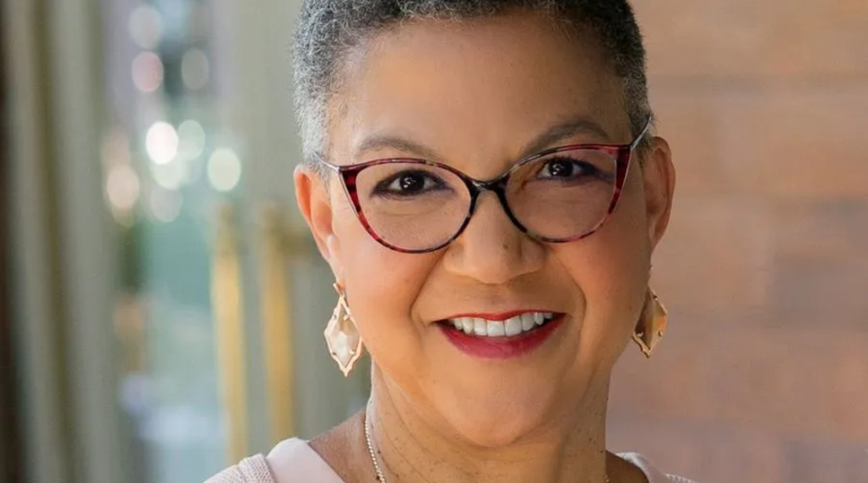 Close-up portrait of a smiling woman with short gray hair, wearing red-framed glasses and gold earrings, standing indoors against a softly blurred background.