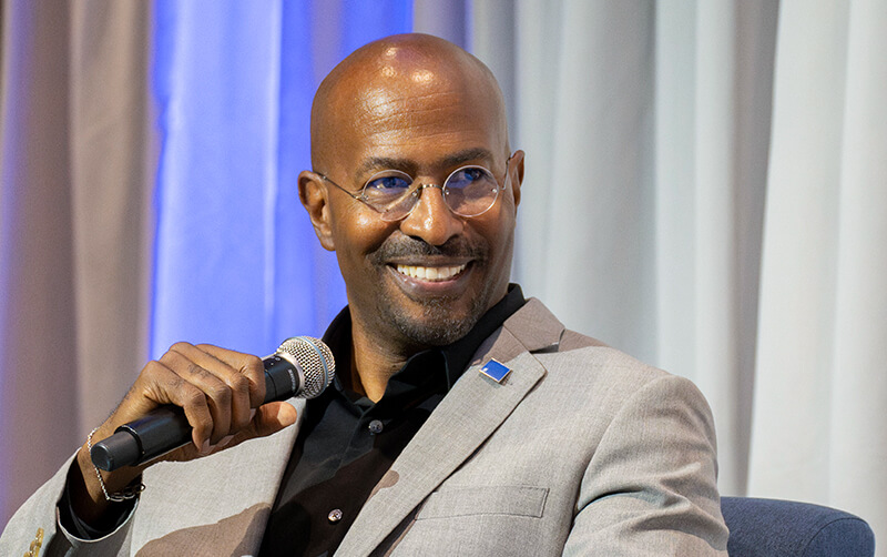 A man in a gray blazer and glasses smiles while holding a microphone during a panel discussion.