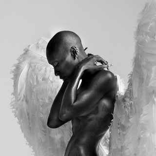 Black-and-white image of a person with a shaved head and feathered wings, posed with arms crossed over their chest against a soft, minimal background.
