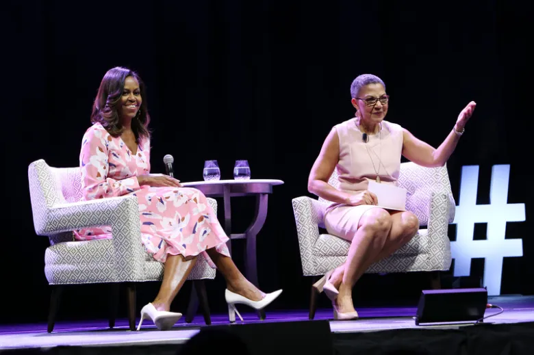 Two women sit on stage in armchairs during a live conversation, holding microphones and speaking to an audience under stage lighting.
