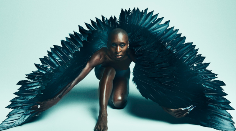 Cynthia Erivo crouched in a dramatic pose wearing large black feathered wings against a minimal studio background.