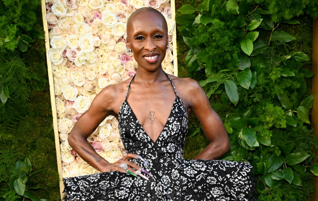 Cynthia Erivo smiling on a red carpet in a black and silver patterned dress, standing in front of a floral backdrop.