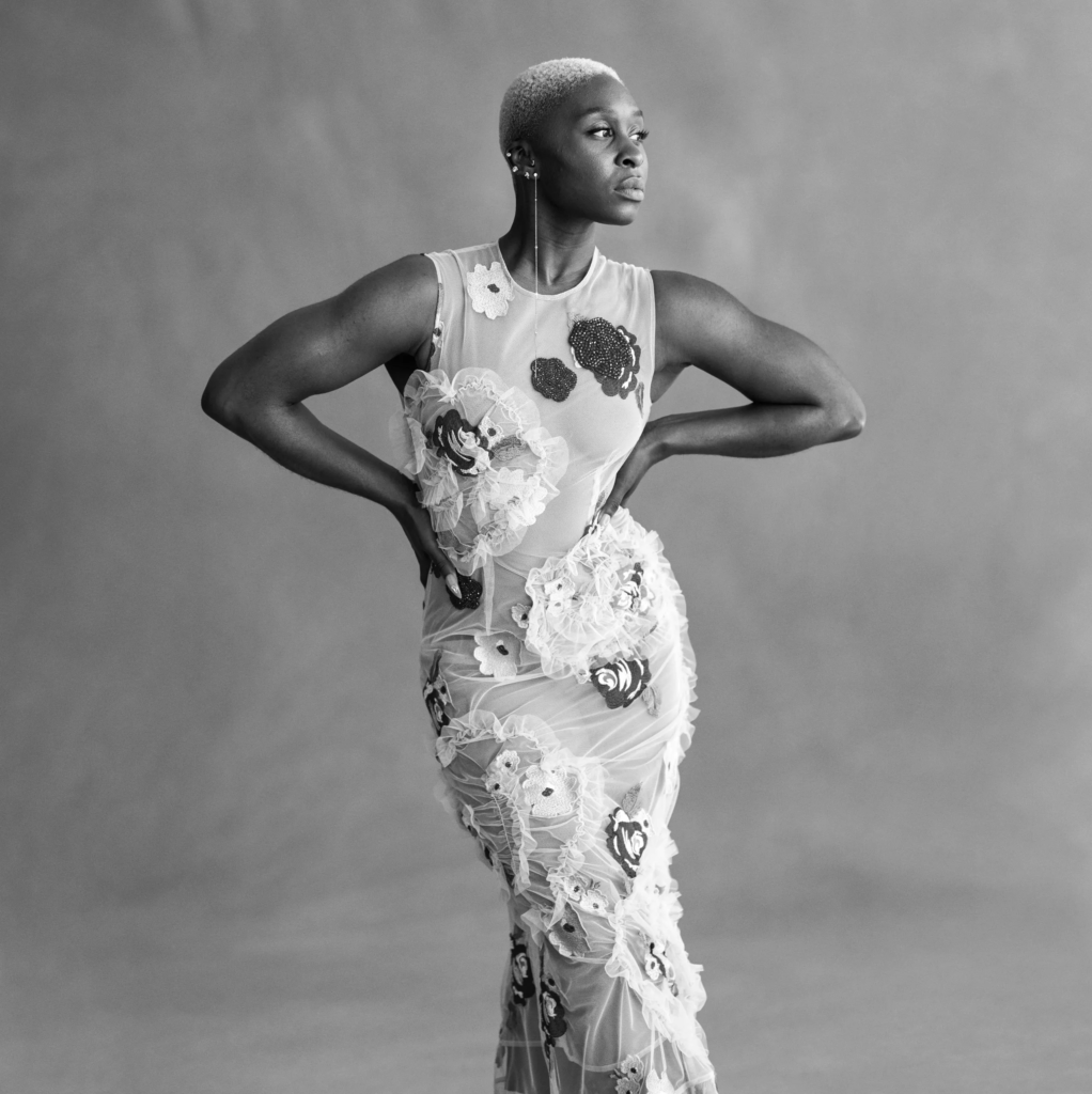 Black-and-white portrait of Cynthia Erivo in a sheer, embellished gown, standing with hands on hips and looking confidently to the side.