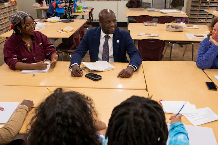 A group sits around tan school tables, looking and talking with each other.