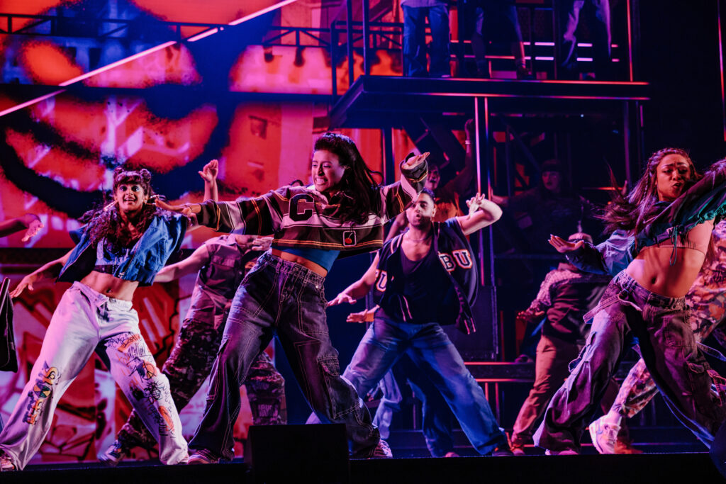 A group of dancers performs a synchronized routine under vivid red and pink lighting, showcasing expressive movement and street-style choreography.
