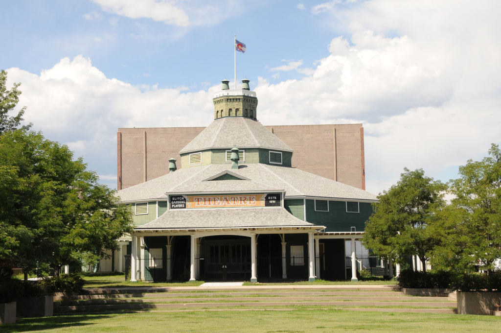 Large historic structure sits on a lawn with green trees on either side of it.