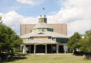 Large historic structure sits on a lawn with green trees on either side of it.