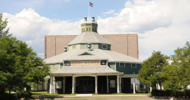 Large historic structure sits on a lawn with green trees on either side of it.