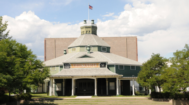 Large historic structure sits on a lawn with green trees on either side of it.