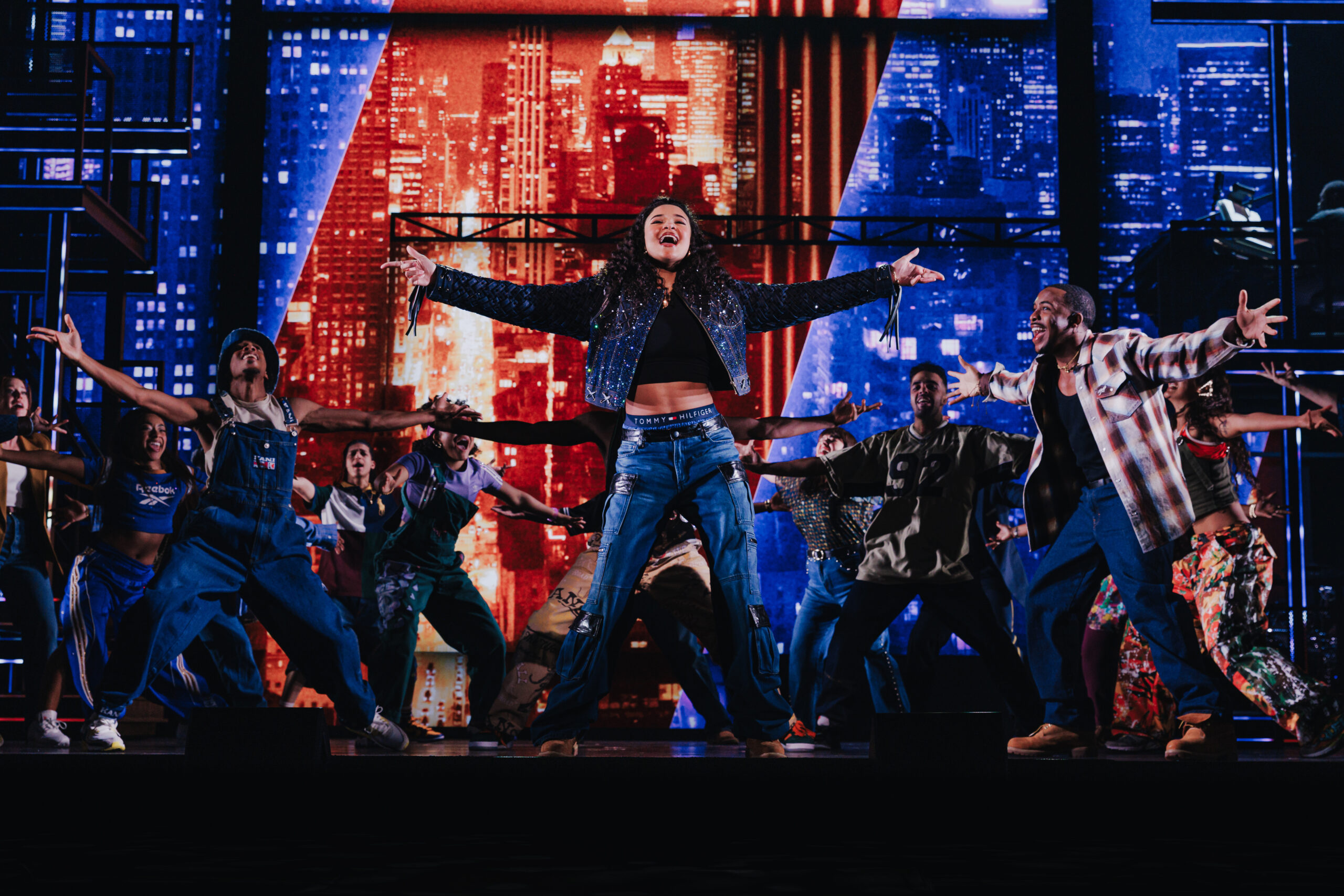 Ali stands center stage with arms outstretched, surrounded by a high-energy ensemble in a dynamic dance number, set against a vibrant New York City skyline backdrop.