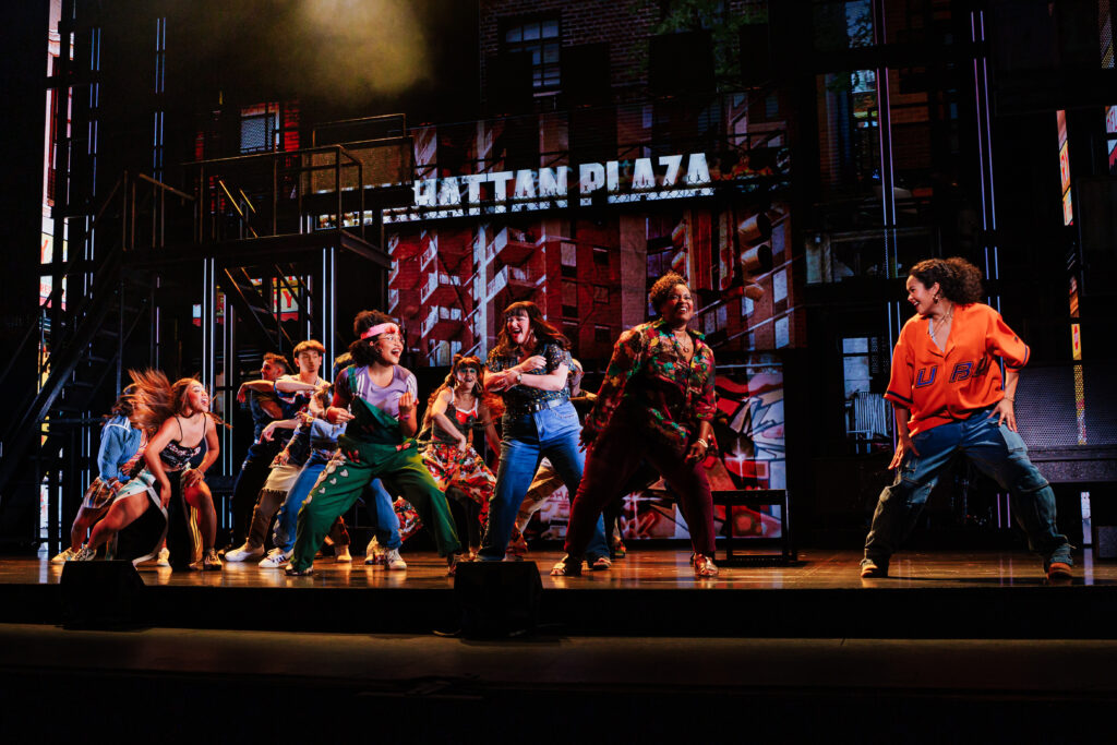 A group of performers dance energetically on a multi-level urban set featuring a “Manhattan Plaza” backdrop, with colorful streetwear costumes and expressive choreography capturing the energy of New York City.