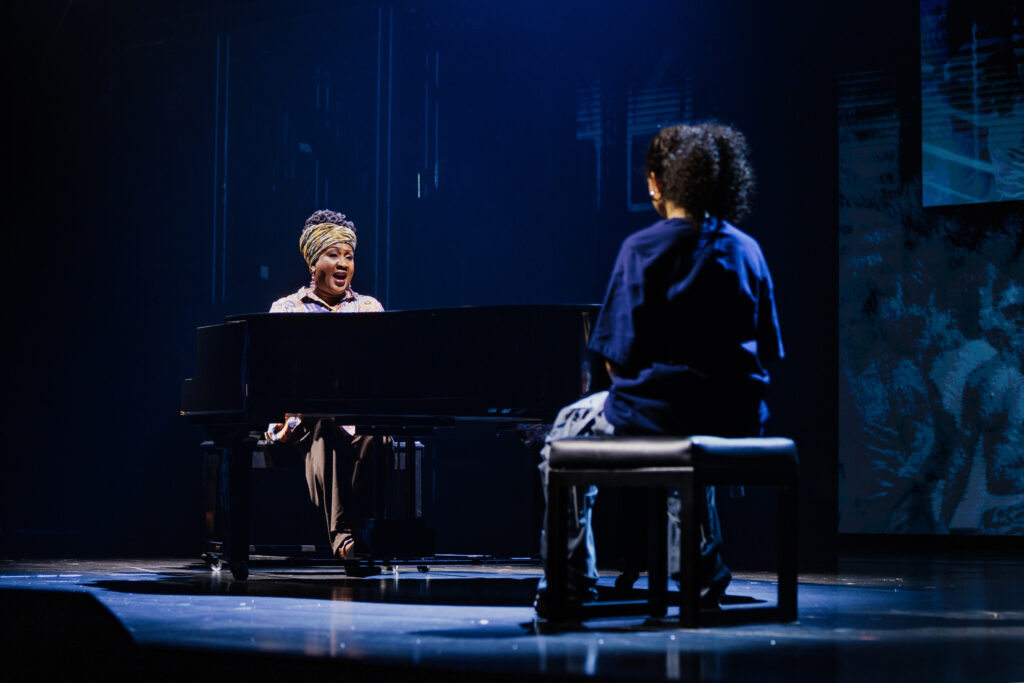 Miss Liza Jane sits at a grand piano, singing passionately while Ali listens nearby, spotlighted in an intimate and emotional moment on a dimly lit stage.