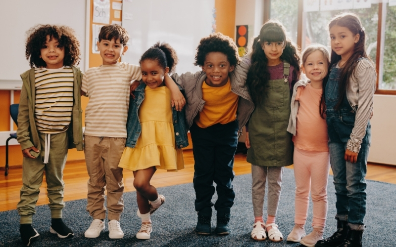 Group of young children standing together in a classroom with their arms around each other.