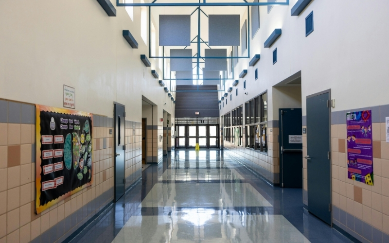 Empty school hallway with blue tile floors, classroom doors, and sunlight coming through large windows.