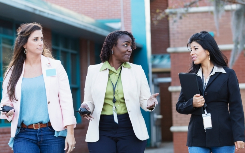 Three professional women walking outside near a school or office building while talking together, holding phones and folders.
