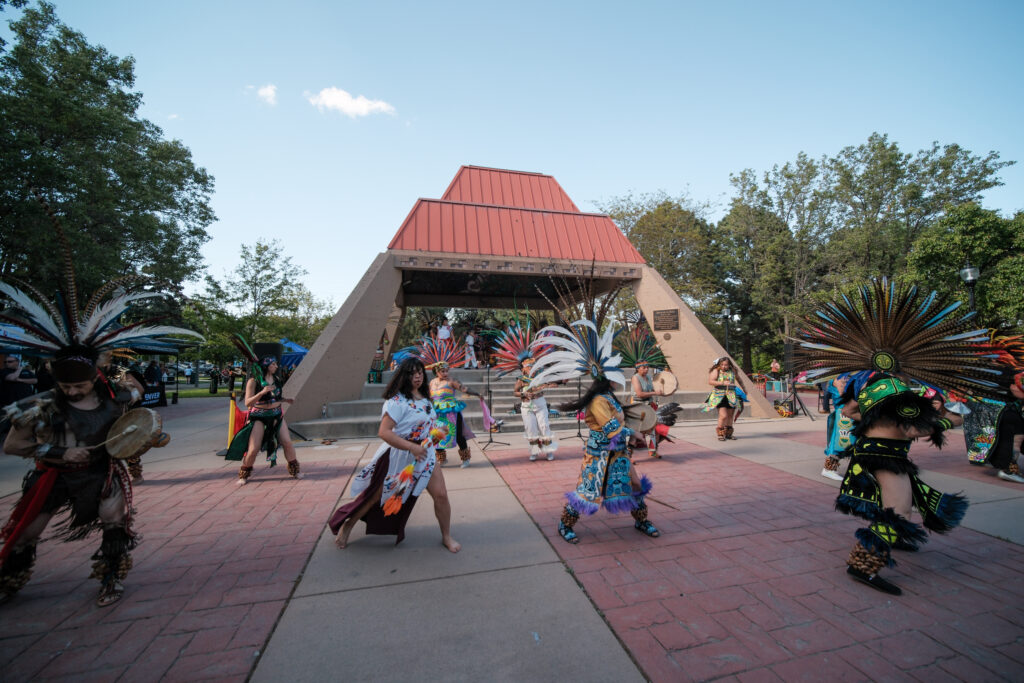 Dancers in colorful traditional attire perform in front of the La Raza Park pavilion during a cultural celebration.