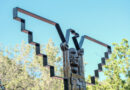 Bronze sculpture featuring two faces and raised arms framed by geometric metal shapes at La Raza Park against a blue sky.