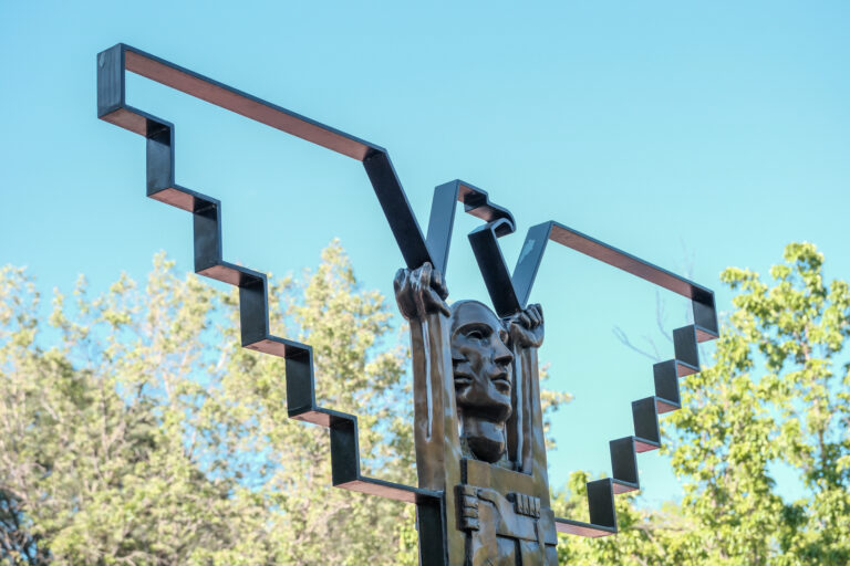 Bronze sculpture featuring two faces and raised arms framed by geometric metal shapes at La Raza Park against a blue sky.