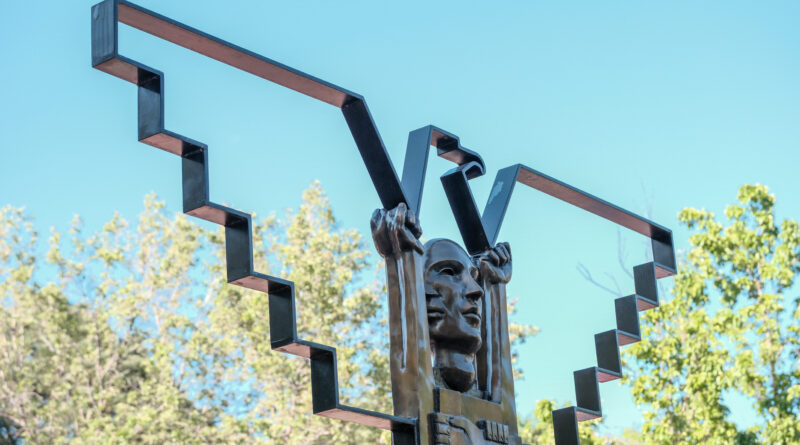 Bronze sculpture featuring two faces and raised arms framed by geometric metal shapes at La Raza Park against a blue sky.