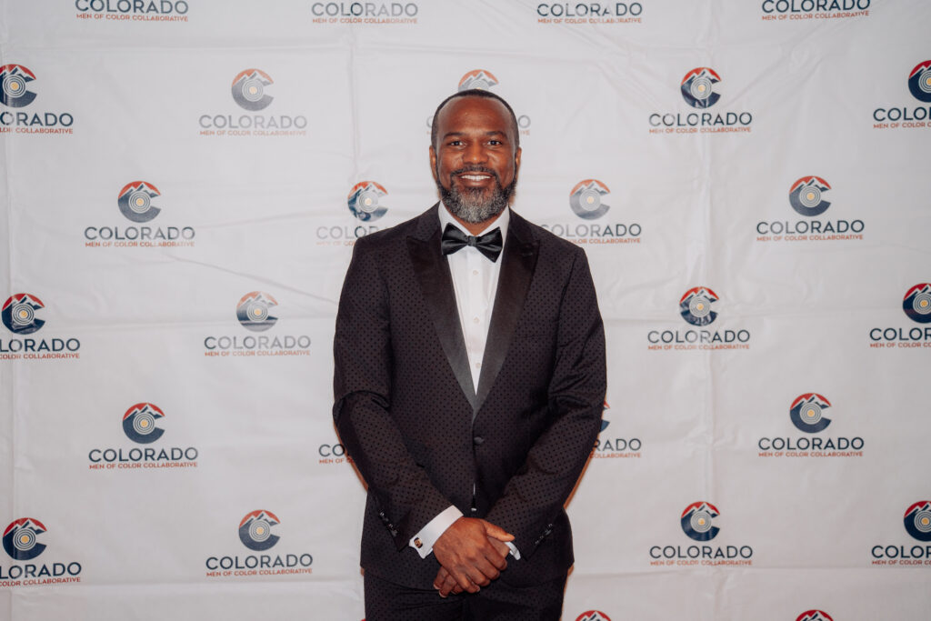 Dr. Fernando Branch poses in a black tuxedo in front of a Colorado Men of Color Collaborative step-and-repeat backdrop during a formal community event.