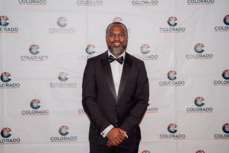 Dr. Fernando Branch poses in a black tuxedo in front of a Colorado Men of Color Collaborative step-and-repeat backdrop during a formal community event.