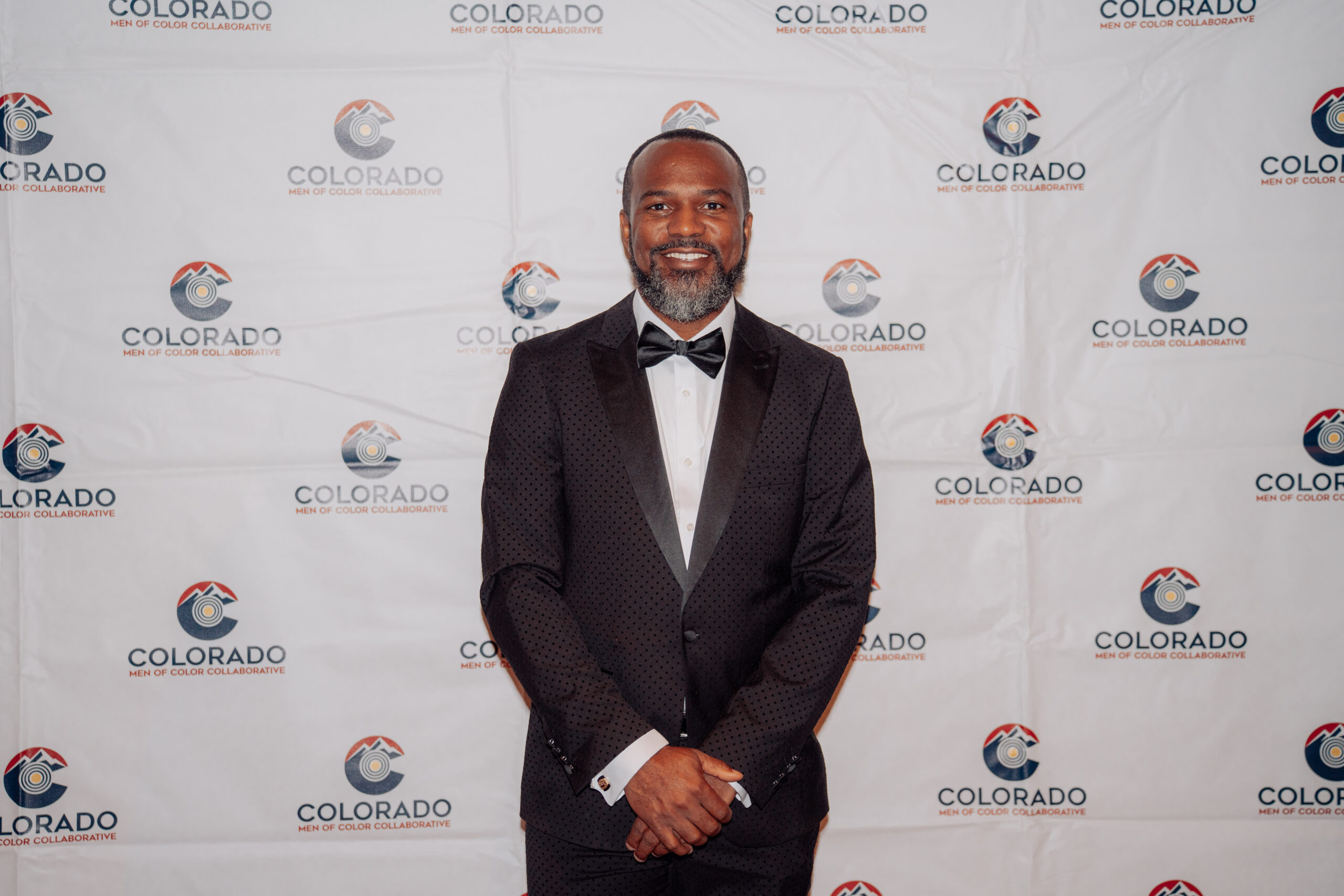 Dr. Fernando Branch poses in a black tuxedo in front of a Colorado Men of Color Collaborative step-and-repeat backdrop during a formal community event.