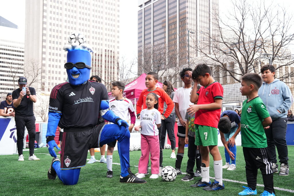 The Colorado Rapids mascot interacts with a group of children on the Street Soccer USA field at Skyline Park, creating a fun and welcoming community soccer experience in downtown Denver.