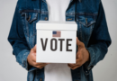 Man wearing dark denim jacket holds white ballot box with the word "Vote" on it