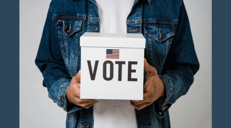 Man wearing dark denim jacket holds white ballot box with the word "Vote" on it