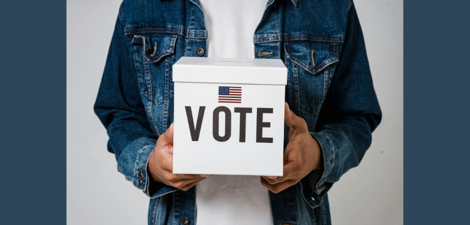 Man wearing dark denim jacket holds white ballot box with the word "Vote" on it