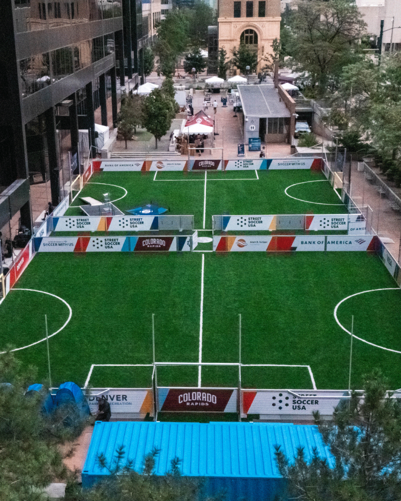 Aerial view of the Street Soccer USA mini-pitch at Skyline Park in downtown Denver, featuring two small-sided soccer fields surrounded by office buildings, trees, and community gathering spaces.